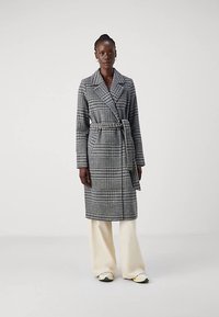 Μακρύ γκρι καρό παλτό με ζώνη στη μέση, φαρδιές πέτο και πλαϊνές τσέπες, συνδυασμένο με κρεμ φαρδιά παντελόνια και αθλητικά παπούτσια.