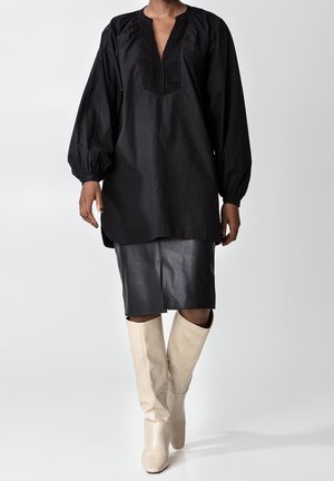 Svart långärmad tunika med en texturerad v-ringning, kombinerad med en svart läderpennkjol och krämfärgade knähöga stövlar.