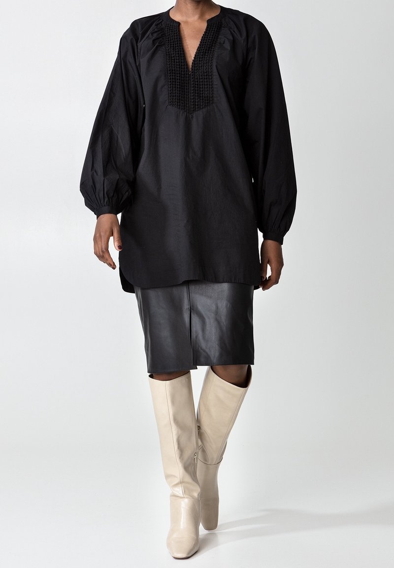 Svart långärmad tunika med en texturerad v-ringning, kombinerad med en svart läderpennkjol och krämfärgade knähöga stövlar.