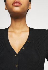 Black ribbed knit garment with a V-neckline, featuring gold buttons along the placket. Smooth texture with fitted silhouette.