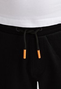 Pantalones deportivos negros con cordón decorado con puntas naranjas, combinados con una camiseta blanca ajustada; tela suave, cintura elástica y detalles plisados.