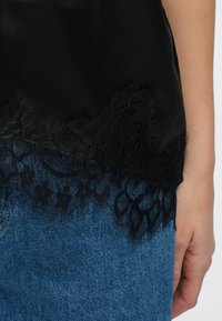 Zwarte satijnen top met een geschulpte kanten rand bij de zoom. Het kant heeft bloemenpatronen en fijne franjes, gecombineerd met blauwe denim.