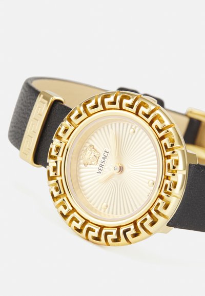 Ceas de aur cu un cadran cu model striat, având o bezel cu design de cheie grecească și o curea din piele texturată neagră. Logo-ul Versace este afișat.