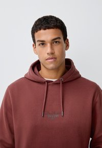 Sweat à capuche bordeaux en tissu doux, avec capuche à cordon de serrage et logo imprimé sur le devant. Texture lisse avec une coupe décontractée.