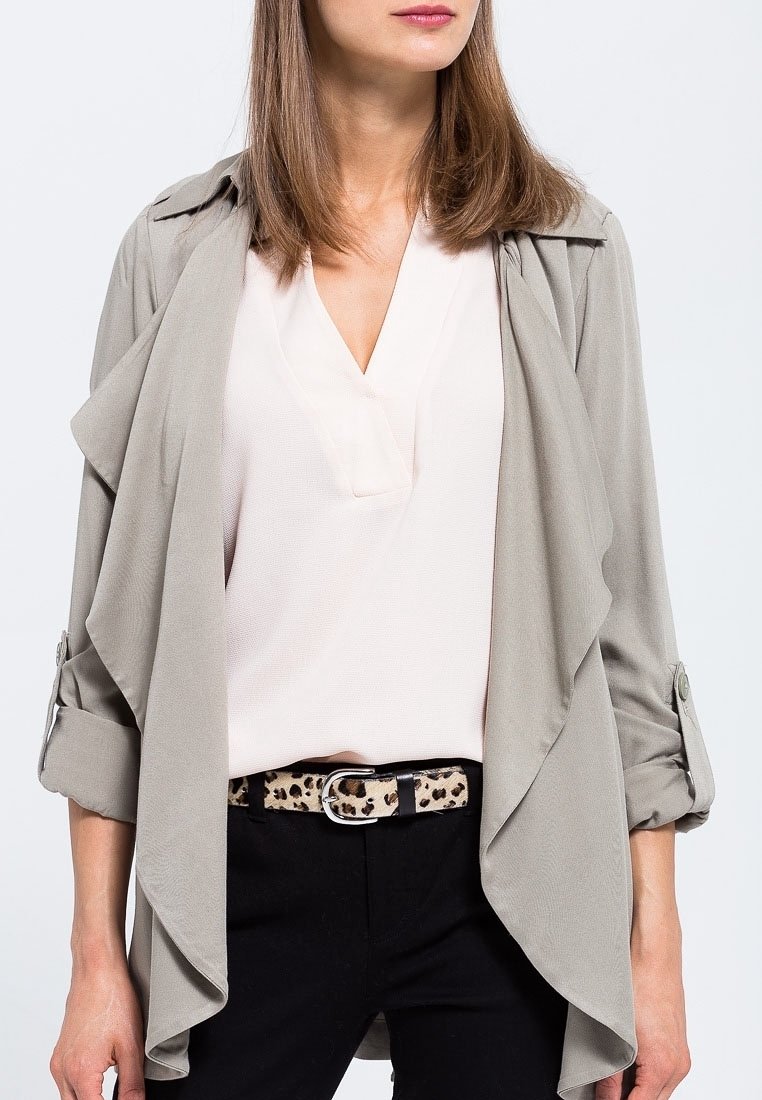 Femme portant une veste drapée beige clair, un chemisier blanc, un pantalon noir et une ceinture imprimé léopard des épaules aux hanches.