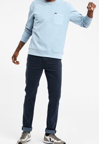 Man med ljusblå långärmad skjorta, mörkblå uppviktbyxor och grå sneakers står mot en enkel vit bakgrund.