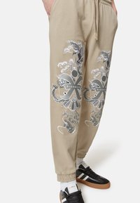 Pantaloni jogger beige con vita regolabile, caratterizzati da stampe grafiche nere e grigie e polsini elasticizzati. Abbinati a sneakers nere.