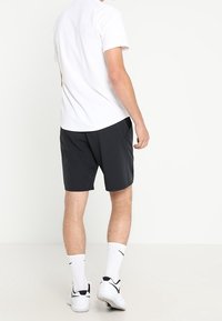 Camisa blanca de manga corta y pantalones cortos negros, con una textura suave. Llevada con calcetines blancos y zapatillas deportivas negras, vista desde atrás.