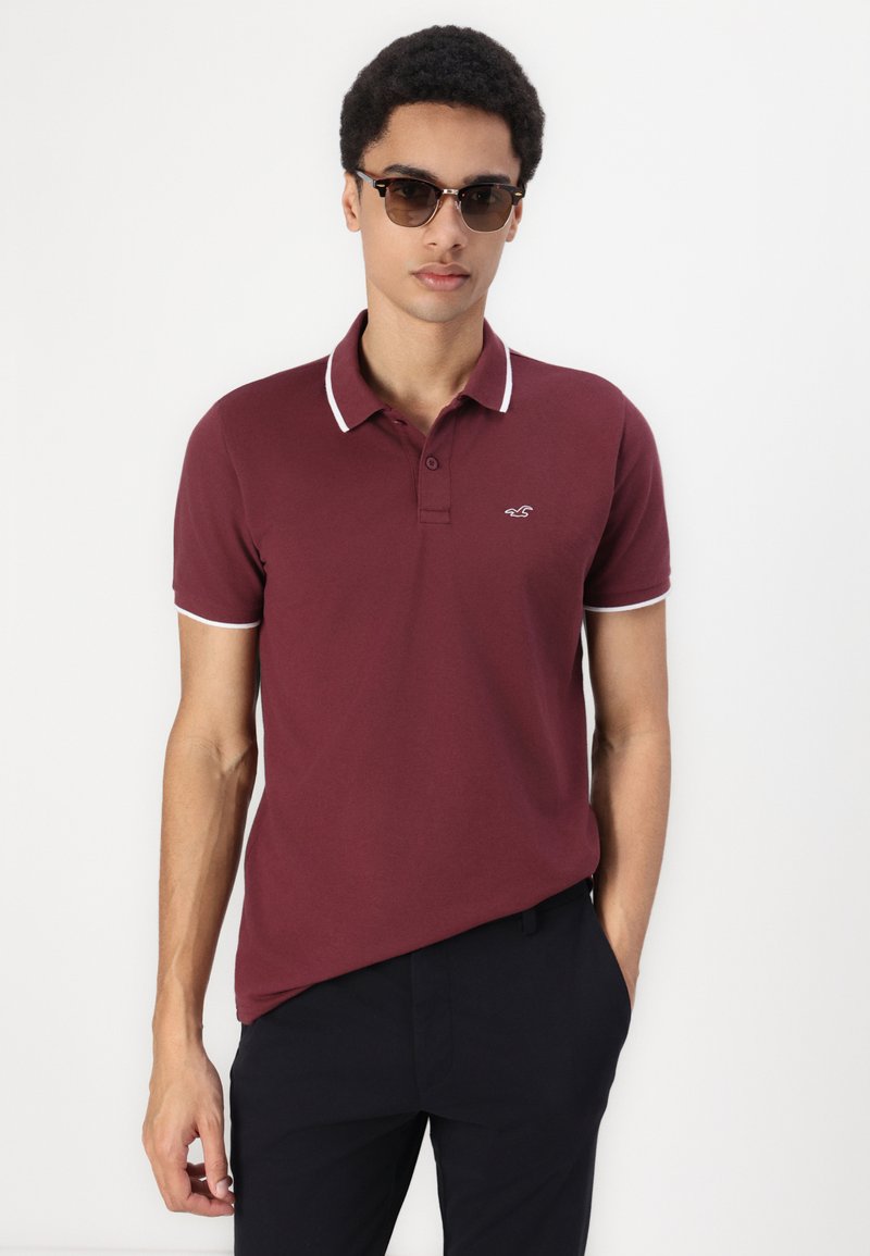Polo de couleur bordeaux avec des bordures blanches au col et aux manches, arborant un petit logo. Le modèle porte des lunettes de soleil noires et un pantalon noir.