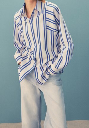 Person wearing a loose white button-up shirt with blue vertical stripes and light blue jeans, standing against a plain blue background.