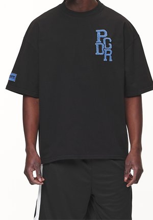 Czarny oversize t-shirt z niebieskimi haftowanymi literami "PGDR" na piersi oraz niebieską naszywką na lewym rękawie, noszony przez osobę.