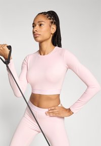 Φωτεινό ροζ cropped μπλουζάκι με μακριά μανίκια και αντιστοιχισμένη ζώνη μέσης με λογότυπο. Το μοντέλο κρατά μια μαύρη ελαστική ταινία, αναδεικνύοντας το εφαρμοστό σχέδιο.