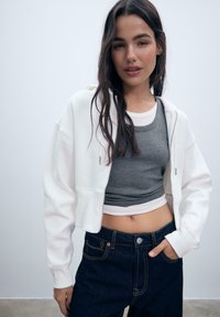 Strihaná biela mikina na zips, šedý priliehavý tielko, tmavomodré nohavice s vysokým pásom a oranžovým prešívaním, slim fit, hladká textúra.