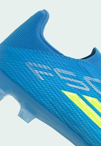 Chaussure de football bleue avec empeigne texturée, texte blanc "F50" et bandes jaunes sur le côté, avec des crampons en plastique transparent sur la semelle.