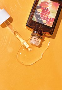 Botella de vidrio marrón con un cuentagotas, que contiene suero amarillo, derramándose sobre un fondo naranja. La etiqueta presenta texto y un gráfico de fruta.