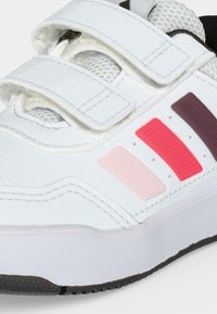 Baskets blanches avec trois bandes velcro et trois bandes colorées en rose clair, rouge et violet foncé sur le côté, avec une semelle noire texturée.