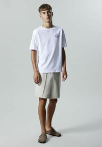 Camiseta de algodón blanca con un diseño gráfico en el pecho, combinada con pantalones cortos beige y zapatillas gris de tipo slip-on. Un atuendo simple y casual.