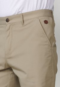 Pantaloni be age dintr-un material neted, cu fermoar la brâu, închidere cu nasture și o mică etichetă logo maro pe buzunarul din față.