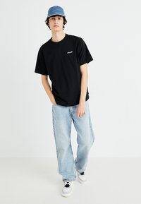 Mustat puuvillateepaidat, joissa on lyhyet hihat ja valkoinen "Levi's"-logo, yhdistettynä vaaleansinisiin farkkuihin ja valkoisiin lenkkikenkiin, joissa on mustia yksityiskohtia.