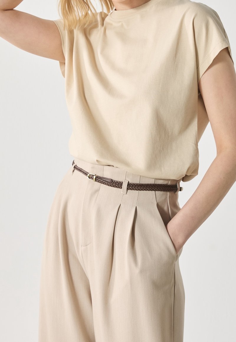 Personne portant un haut beige à manches courtes et un pantalon beige taille haute avec des plis et une fine ceinture tressée marron, main dans la poche.