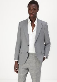 Grauer maßgeschneiderter Blazer mit Reverskragen, zwei Knöpfen und frontalen Taschen; getragen über einem weißen Hemd und gemusterten grauen Hosen.