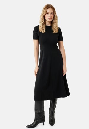 Frau mit welligem blondem Haar, die ein schwarzes Midi-Kleid mit kurzen Ärmeln und schwarze kniehohe Stiefel mit spitzer Spitze trägt, steht vor weißem Hintergrund.