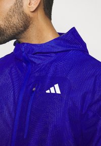 Homme portant une veste à capuche bleu vif avec une poche poitrine zippée et un logo de marque stylisé blanc sur le côté gauche.