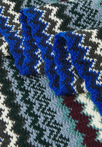Adīta sega ar houndstooth rakstu melnā, baltā, zaļā, zilā un burgundijas krāsā. Strukturēti maliņas ar zilu viļņveida apdari.