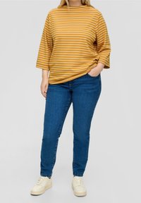 Geel-wit gestreept bovenstuk met een losse pasvorm en driekwartmouwen, gecombineerd met een blauwe spijkerbroek en gebroken-witte sneakers. Gladde stoftextuur.