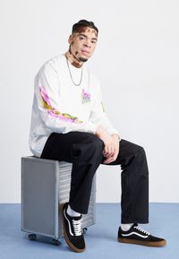 Mladý muž s vitiligem, oblečený v bílém grafickém svetru, černých kalhotách a černých teniskách, sedí na kovové krabici na kolečkách na modré podlaze.