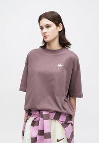 Mlada ženska z temnimi lasmi, oblečena v ohlapen mauve majico z belim logotipom, z večbarvno roza in rjavo jopico, vezano okoli pasu, ter belimi kratkimi hlačami.