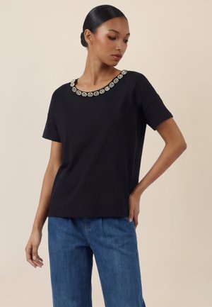 Nainen, jolla on tummat hiukset matalalla nutturalla, musta lyhythihainen paita, jossa on strassikivillä koristeltu pääntie, ja siniset farkut, seisoo toisella kädellä lantiolla.