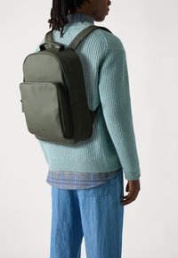 Sac à dos en cuir vert foncé avec une texture lisse, poche avant zippée et bretelles réglables, porté par-dessus un pull en tricot bleu clair.