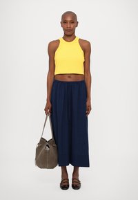 Geel geribbelde crop top, marineblauwe wijde broek, grijze leren handtas en zwarte schoenen. Model staat rechtop tegen een witte achtergrond.