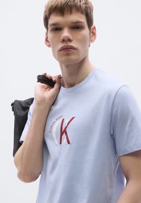 Ανοιχτό γαλάζιο βαμβακερό μπλουζάκι με κόκκινο και λευκό λογότυπο «Calvin Klein». Το μοντέλο κρατά μια μαύρη τσάντα με φερμουάρ.