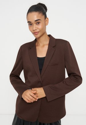 Joven mujer con el cabello oscuro recogido en un moño, lleva una blazer marrón sobre una camiseta negra, de pie con las manos entrelazadas frente a un fondo liso.