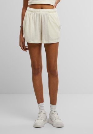 Kvinde iført cremefarvede teksturerede shorts, hvide sneakers og hvide crew-sokker mod en ensfarvet lys baggrund.