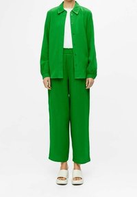 Chemise-veston texturée verte et pantalon à jambes larges, associés à un t-shirt blanc et des sandales compensées de couleur claire.