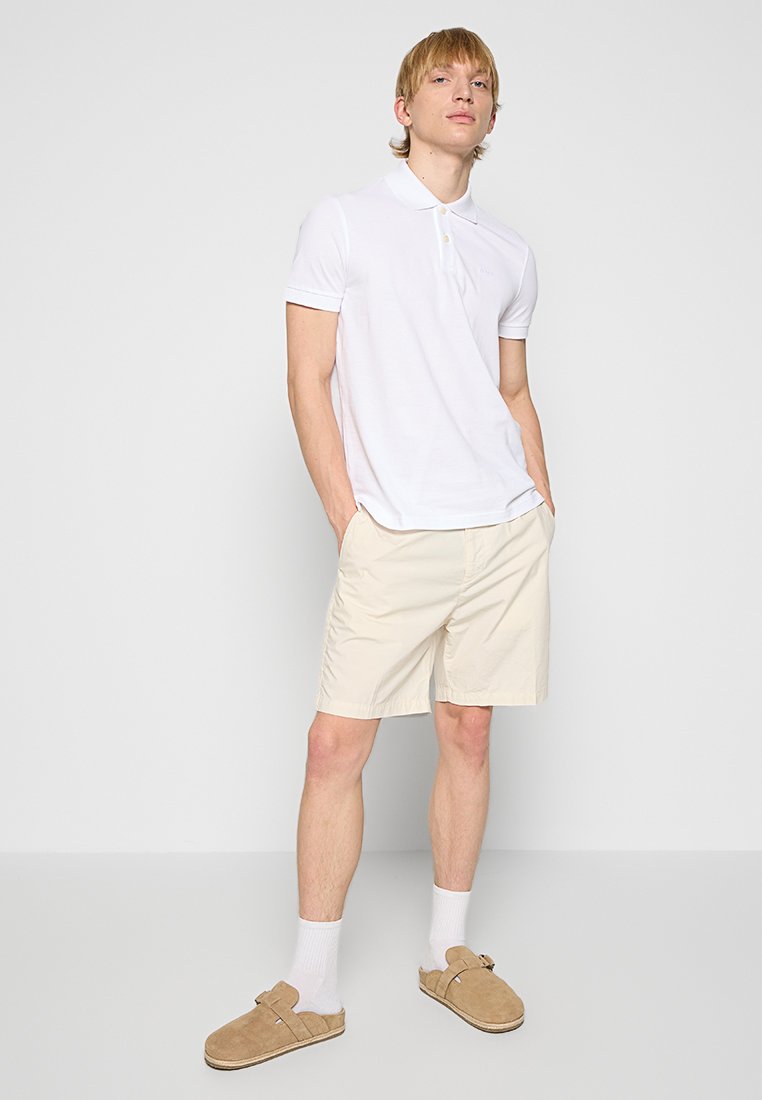 Polo blanc, shorts beiges et sabots en cuir beige. Le polo présente un col et une coupe élégante, tandis que les shorts ont une texture lisse et légère.
