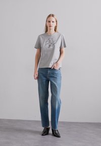 T-shirt in cotone grigio con un sottile design grafico, abbinato a jeans di denim blu chiaro. Il modello indossa scarpe con tacco nere. Sfondo semplice.