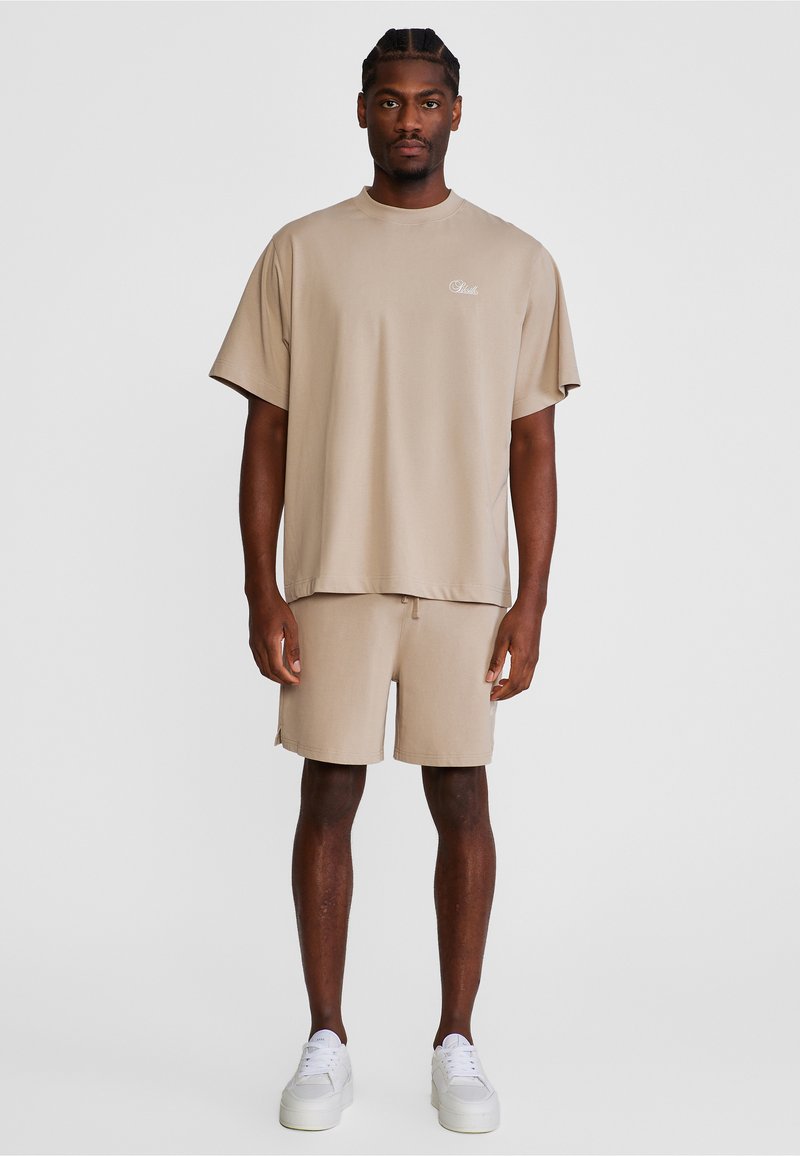 Άνδρας που στέκεται φορώντας μπεζ oversized μπλουζάκι και ταιριαστά σορτς με λευκά αθλητικά παπούτσια μπροστά σε απλό ανοιχτό φόντο.