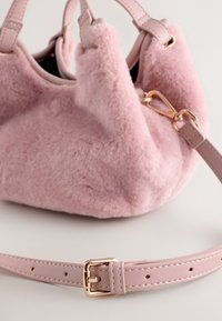 Roosa kunstkarvaga käekott ümara kujuga, nahast sangade ja eemaldatava nahast rihmaga. Kaunistatud kullast detailidega ja sileda tekstuuriga.