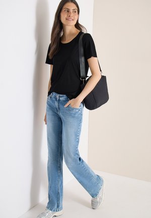 Jeune femme en t-shirt noir et jean bleu clair s'appuie contre un mur, portant un sac à bandoulière noir et des baskets blanches.