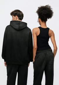 Μαύρο φαρδύ hoodie με υφή επιφάνειας, συνδυασμένο με εφαρμοστή μαύρη μπλούζα χωρίς μανίκια και ταιριαστά στενά παντελόνια με στενή γραμμή. Μινιμαλιστικός σχεδιασμός, χωρίς ορατά μοτίβα.