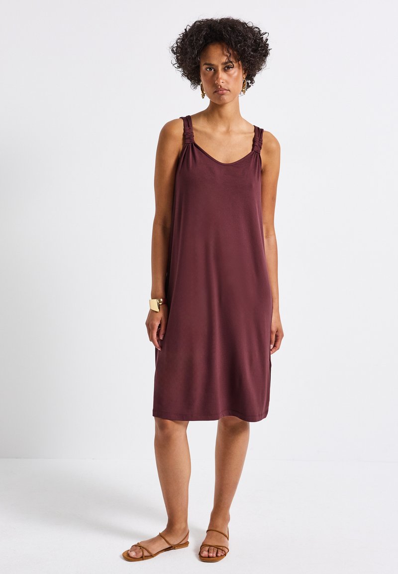Burgundy ärmlös klänning med elastiska axelband, mjukt tyg, avslappnad passform och medellång design. Buren med minimaliska sandaler.