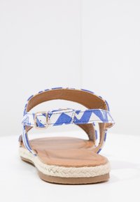 Sandal i blå og hvidmønstrede stoffer med justerbar rem, vævet reb-sål og en glat brun fodseng.