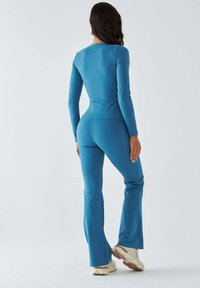 Kombinezon na całej długości z długim rękawem, wykonany z jednolitej niebieskiej tkaniny, z dopasowanym topem i rozkloszowanymi spodniami, w połączeniu z jasnymi butami sportowymi.