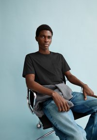 Zwart t-shirt met korte mouwen en een borstzak, gecombineerd met lichtblauwe spijkerbroek. Een grijze fleece is over de schoot gedrapeerd. Chroom bureaustoel.