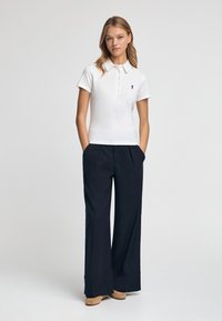 Biała koszulka polo z krótkim rękawem i trzema guzikami, w połączeniu z szerokimi granatowymi spodniami i beżowym obuwiem. Prosty logo na koszulce.