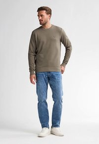 Mörk olivgrön crewneck-tröja i mjukt, stretchigt tyg med en liten logotyp. Kombinerad med ljusblå denimjeans och vita sneakers.
